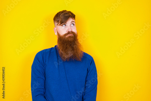 Young handsome red haired bearded man standing against yellow wall has worried face looking up lips together, being upset thinking about something important, keeps hands down.