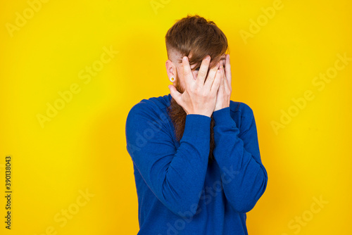 Young handsome red haired bearded man standing against yellow wall covering face with hands and peering out with one eye between fingers. Scared from something or someone. © Jihan