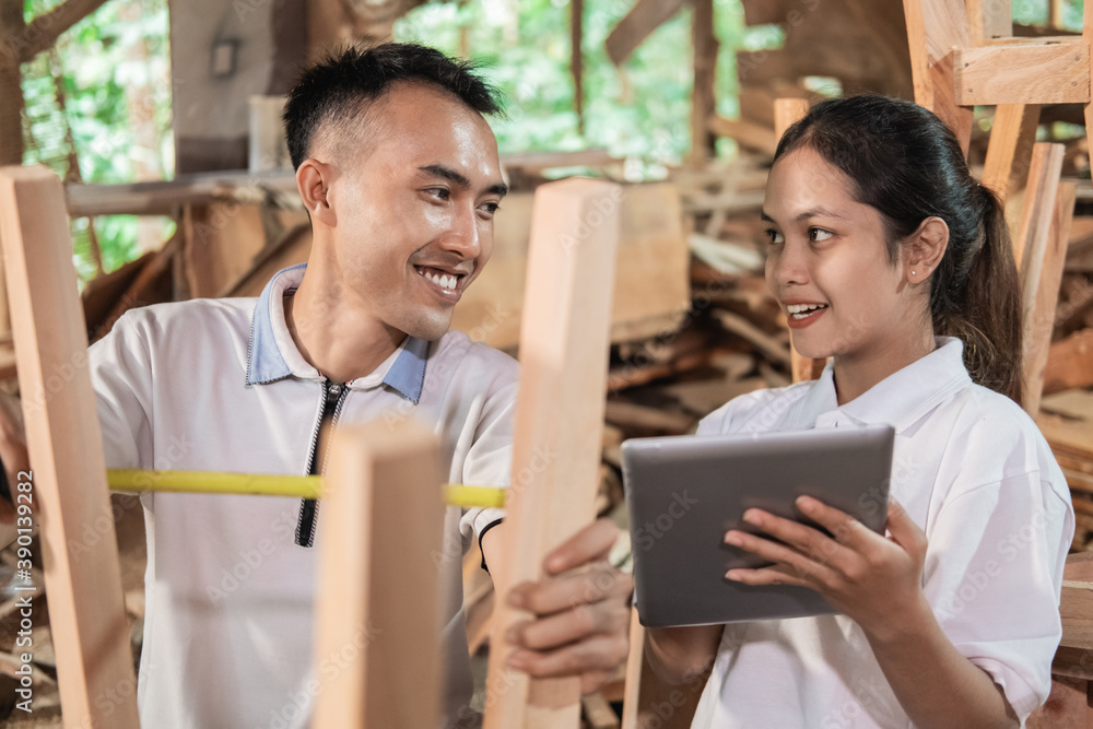 a business woman uses a tablet when she sees a carpenter measuring wood for furniture a business woman uses a tablet when she sees a carpenter measuring wood for furniture