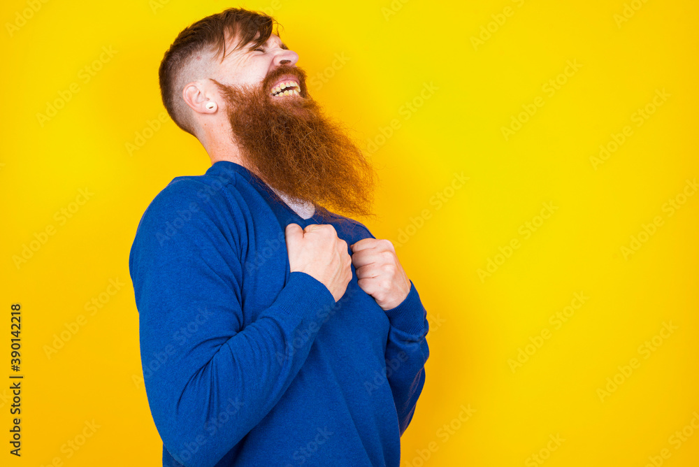 Young handsome red haired bearded man standing against yellow wall excited and glad to achieve victory, clenches fists, screams in excitement with closed eyes,successful person. Young handsome red haired bearded man standing against yellow wall excited and glad to achieve victory, clenches fists, screams in excitement with closed eyes,successful person.