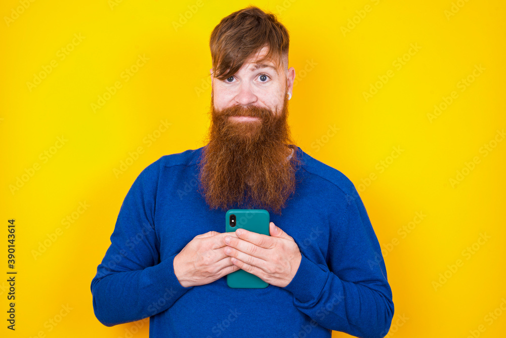 Smiling Young handsome red haired bearded man standing against yellow wall friendly and happily holding mobile phone taking selfie in mirror. Smiling Young handsome red haired bearded man standing against yellow wall friendly and happily holding mobile phone taking selfie in mirror.