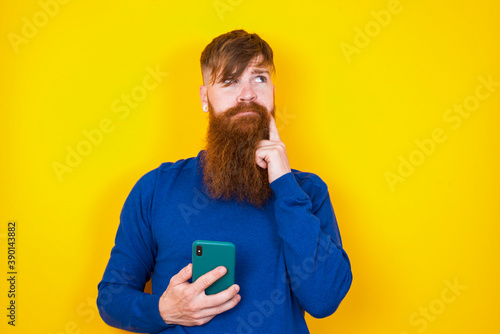 Photos Image of a thinking dreaming Young handsome red haired bearded man standing against yellow wall using mobile phone and holding hand on face