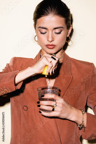 young woman in brown blazer squeezing lemon in glass with alcohol cocktail on white