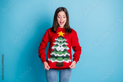 Photo portrait of surprised girl pulling ugly christmas sweater with hands smiling looking down isolated on pastel light blue colored background
