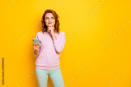 Photo of pretty lady hold telephone hand on chin look empty space think over new smart phone buying wear pink pullover green pants isolated yellow color background