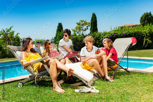 Group of happy teenage boys and girls drink cold cola sit near the pool on chaise longue in the garden talk © Sergey Novikov