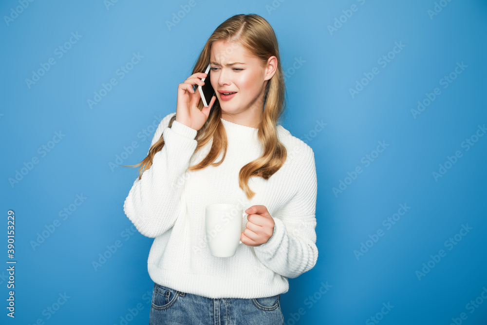 confused blonde woman in sweater talking on smartphone with mug isolated on blue background confused blonde woman in sweater talking on smartphone with mug isolated on blue background