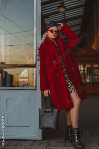 Outdoor fashion portrait of elegant woman wearing red faux fur teddy bear coat, sunglasses, leather beret, ankle boots, holding tote bag, posing in street of city. Autumn, winter trendy outfit. 