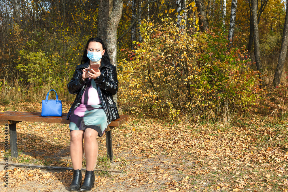 pregnant woman in the fall on a park bench in a protective mask works in a smartphone. pregnant woman in the fall on a park bench in a protective mask works in a smartphone.