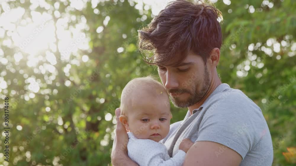 Loving father cuddling and kissing baby daughter as he holds her outdoors in park or garden against flaring sun - shot in slow motion Loving father cuddling and kissing baby daughter as he holds her outdoors in park or garden against flaring sun - shot in slow motion