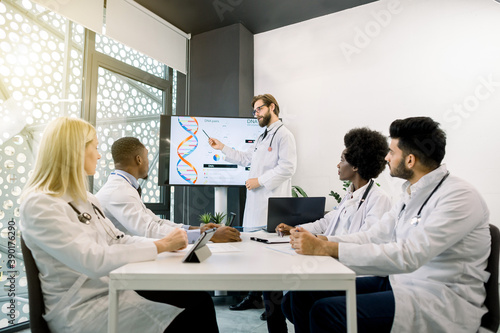 Team of multiethnic doctors in conference room, looking at their colleague, young Caucasian man doctor or biochemist, showing on the screen some biochemical formulas. Medicine, science concept