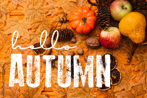 top view of autumnal decoration and food scattered from wicker basket near hello autumn lettering on golden foliage