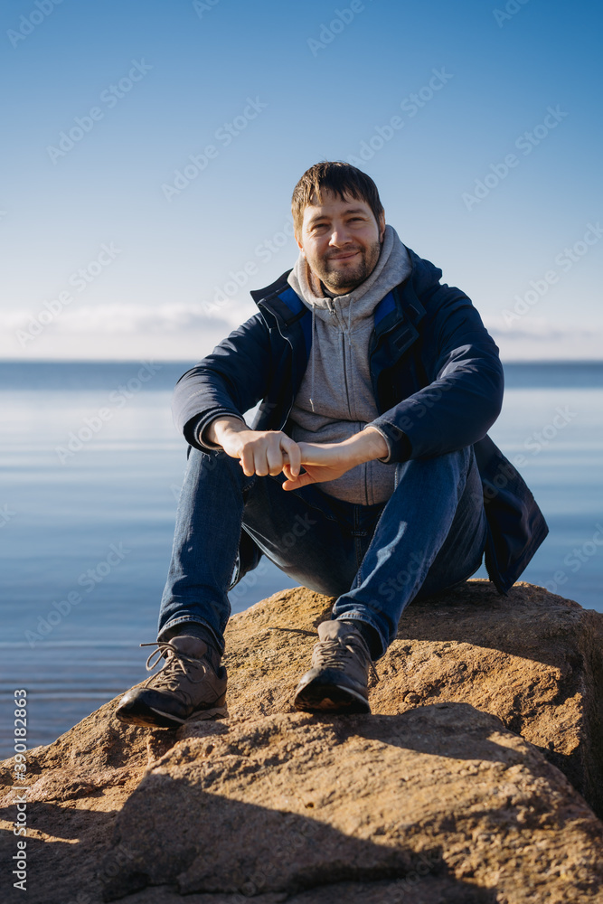 young caucasian man squinting into the sun on warm autumn day sitting on big rock on the shore of finland gulf.