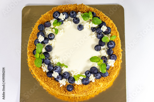 Homemade honey cake with sour cream on a golden plate and white table with blueberries and leaves of mint. Delicious Russian Cuisine.