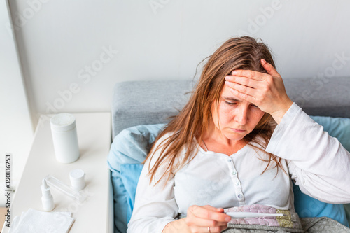 Sick woman checking her temperature with headache and fever lying under the blanket. Sick woman staying in bed with temperature durong coronavirus pandemic. photo