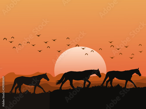 The silhouette of a herd of horses on the hills at sunset has mountains and orange sky as background