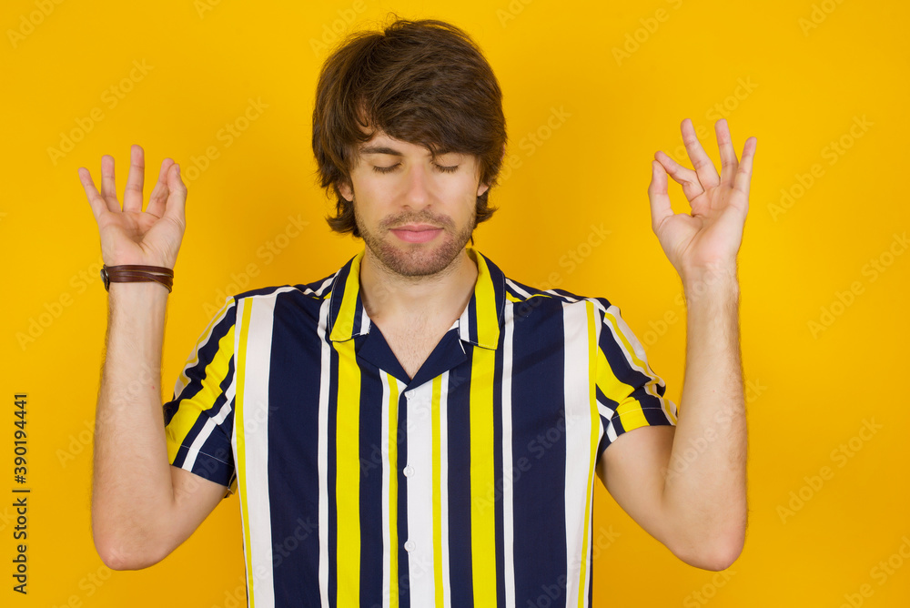 Young handsome Caucasian man, wearing stripped shirt standing against yellow wall doing yoga, keeping eyes closed, holding fingers in mudra gesture. Meditation, religion and spiritual practices.