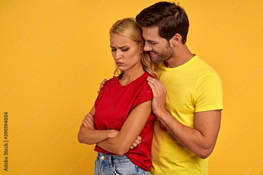 Naklejka premium Offended smiling young couple friends in colored t-shirts posing isolated on yellow background studio portrait. People lifestyle concept. Mock up copy space. Holding hands crossed, hugging.