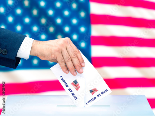 A man's hand voting by mail in the US presidential election with the US flag behind it. Selective focus © javidestock