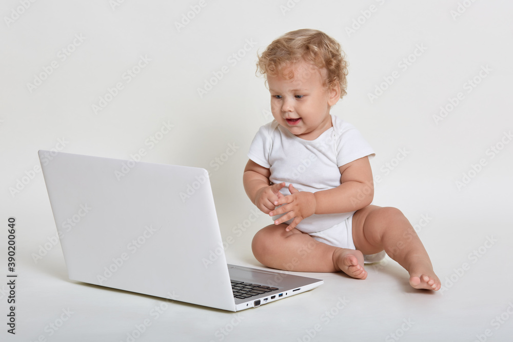 Little blond curly haired male wearing body suit, sitting on floor with lap top, looking at gadget screen with curious facial expression, posing isolated over white background. Little blond curly haired male wearing body suit, sitting on floor with lap top, looking at gadget screen with curious facial expression, posing isolated over white background.