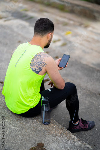 Male using sport training app in phone. Athletic guy sitting on stairs with bottle of water, back view
