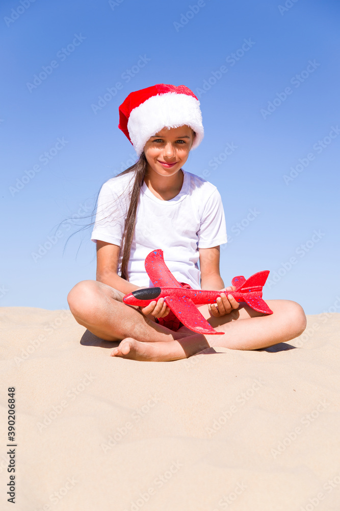 Obraz premium Christmas celebration vacations on a tropical beach: Happy cute little girl wearing a red Santa hat sitting on the sandy beach by the sea and holding red toy plane on her hands on sunny day, vertical