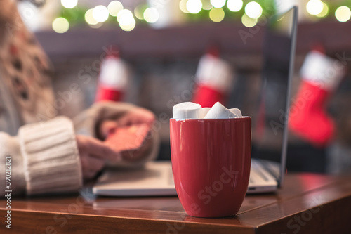 Woman  holding credit card using laptop for making order sitting at table with cup of hot cocoa and marshmallow at christmas fireplace with decoration of light bulbs. Close up.