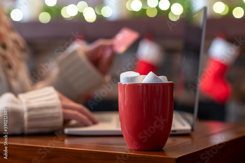 Woman in sweater holding credit card using laptop for making order sitting at table near christmas fireplace with decoration of light bulbs. Online booking, reservation. Travel concept