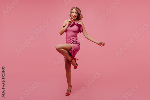 Full-length portrait of excited curly girl standing on one leg on pink background. Stylish glamorous woman dancing with sincere smile