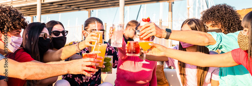 Group of happy friends with face mask drinking and toasting coloured cocktail at bar restaurant - Friendship concept with young people having fun together at pub - New normal lifestyle photo