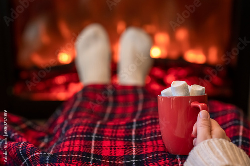 Woman with cup of hot cocoa and marshmallow warming legs in winter white socks near fireplace flame, covered christmas plaid