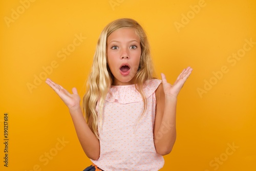 Beautiful Caucasian young girl standing against yellow background shocked rising arms looking to the camera with stunned expression. 