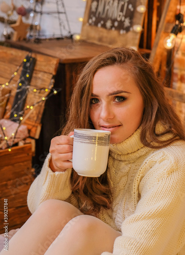 Portrait of young woman with cup of cocoa on bed at home. Minimalist christmas cozy decor on the background. Merry Christmas and Happy Holidays. Blonde, cute, woman in white sweater and warm scandinav photo