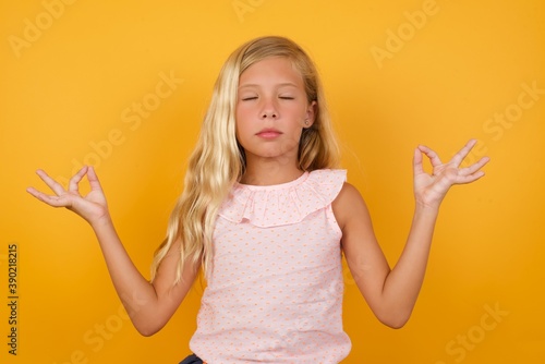 Beautiful Caucasian young girl standing against yellow background doing yoga, keeping eyes closed, holding fingers in mudra gesture. Meditation, religion and spiritual practices.