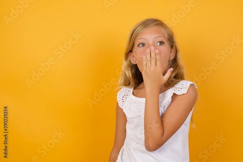 Oh! I think I said it! Close up portrait Beautiful Caucasian young girl standing against yellow background cover open mouth by hand palm, look at camera with big eyes.