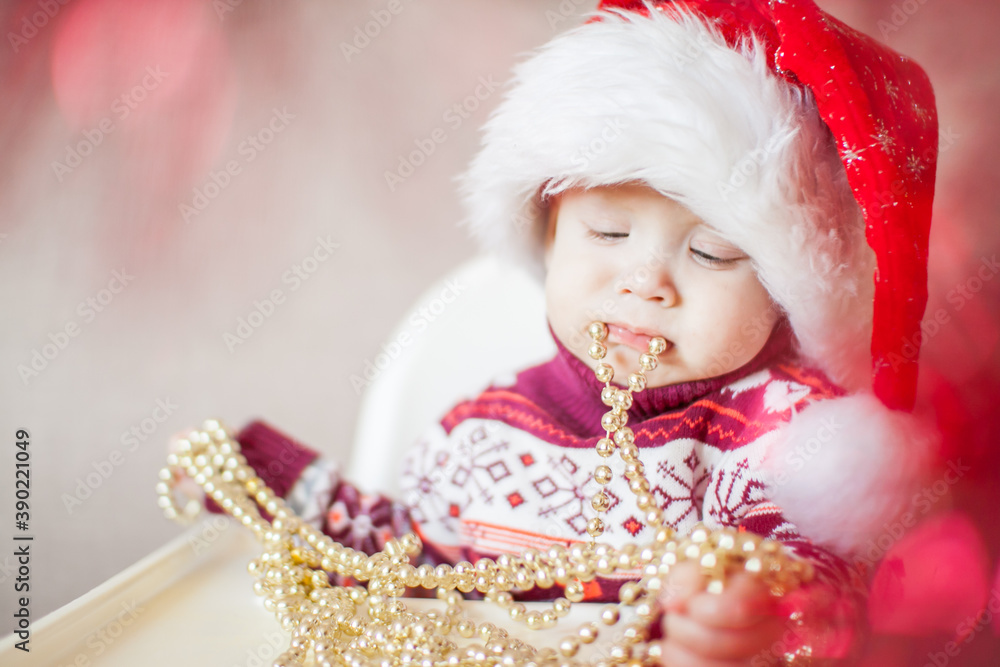 A little baby girl in a New Year's hat of Santa Claus examines and plays with New Year's decorations. A little baby girl in a New Year's hat of Santa Claus examines and plays with New Year's decorations.