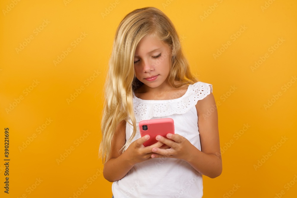 Smiling Beautiful Caucasian little girl standing against yellow background using cell phone, messaging, being happy to text with friends, looking at smartphone. Modern technologies and communication. Smiling Beautiful Caucasian little girl standing against yellow background using cell phone, messaging, being happy to text with friends, looking at smartphone. Modern technologies and communication.