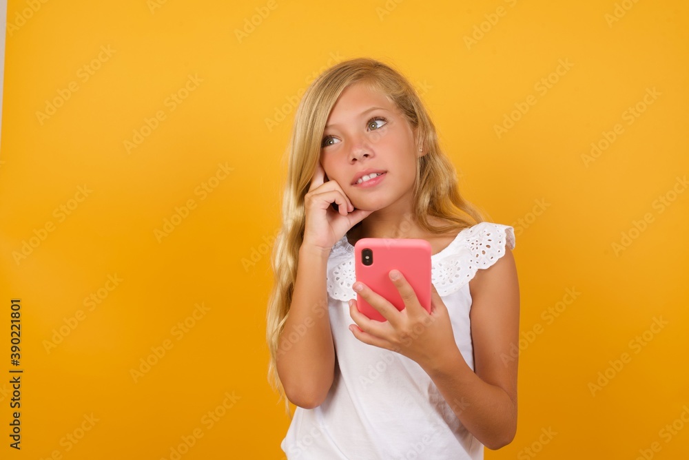 Image of a thinking dreaming Beautiful Caucasian little girl standing against yellow background using mobile phone and holding hand on face. Taking decisions and social media concept. Image of a thinking dreaming Beautiful Caucasian little girl standing against yellow background using mobile phone and holding hand on face. Taking decisions and social media concept.