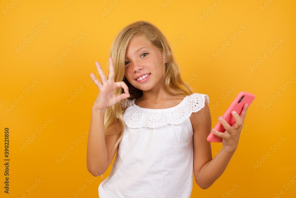 Happy Beautiful Caucasian little girl standing against yellow background sending a message on his smartphone or taking a selfie and making ok sign with his hand. Happy Beautiful Caucasian little girl standing against yellow background sending a message on his smartphone or taking a selfie and making ok sign with his hand.