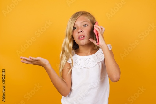 Beautiful Caucasian little girl standing against yellow talking on the phone stressed with hand on face, shocked with shame and surprise face, angry and frustrated. Fear and upset for mistake.