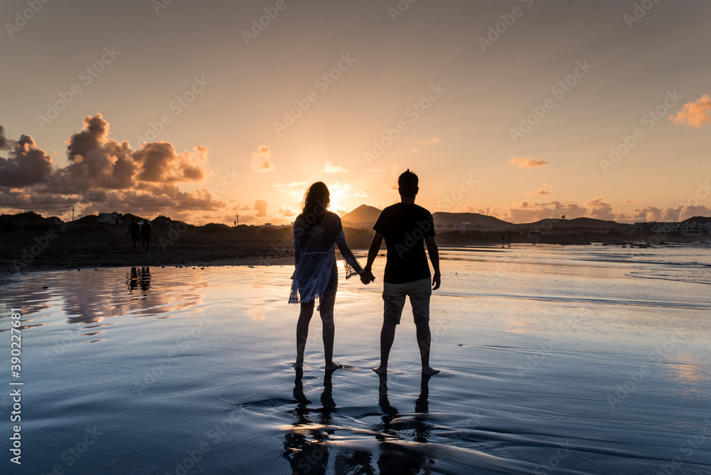 Fototapeta premium Couple from behind holding hands on a beach at sunset.