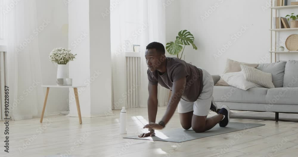 Active black man standing on all fours and practicing swinging legs exercise at home, raising hand and leg