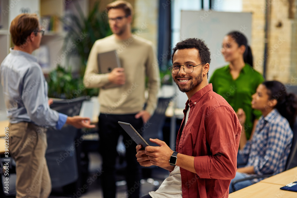 Young cheerful mixed race man holding digital tablet and smiling at camera while working with colleagues in the modern office, selective focus Young cheerful mixed race man holding digital tablet and smiling at camera while working with colleagues in the modern office, selective focus