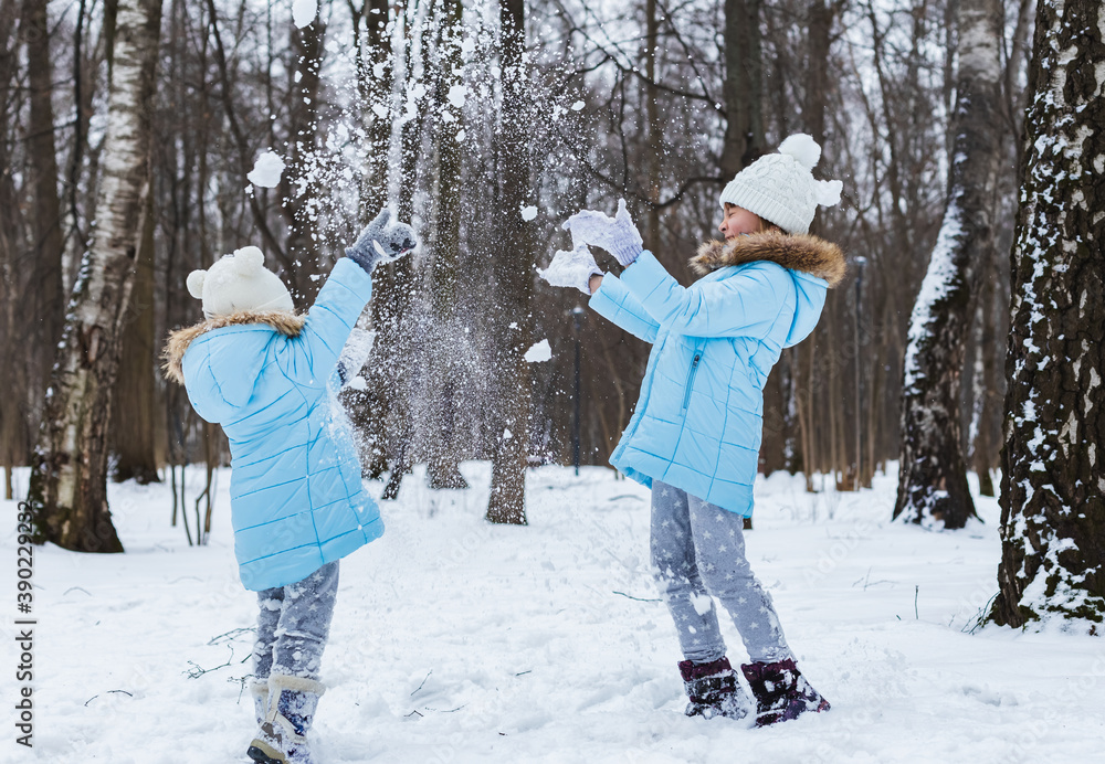 Little girls, sisters are walking, having fun in snowy winter park. Stylish clothes, blue jackets with fur,warm pants with snowflakes,cat-shaped mittens.Family picnic in cold weather.Outdoor activity