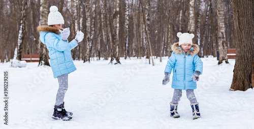 Little girls, sisters are walking, having fun in snowy winter park. Stylish clothes, blue jackets with fur,warm pants with snowflakes,cat-shaped mittens.Family picnic in cold weather.Outdoor activity