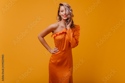 Blithesome white woman in long dress playing with her hair. Indoor photo of good-humoured pretty girl in orange outfit