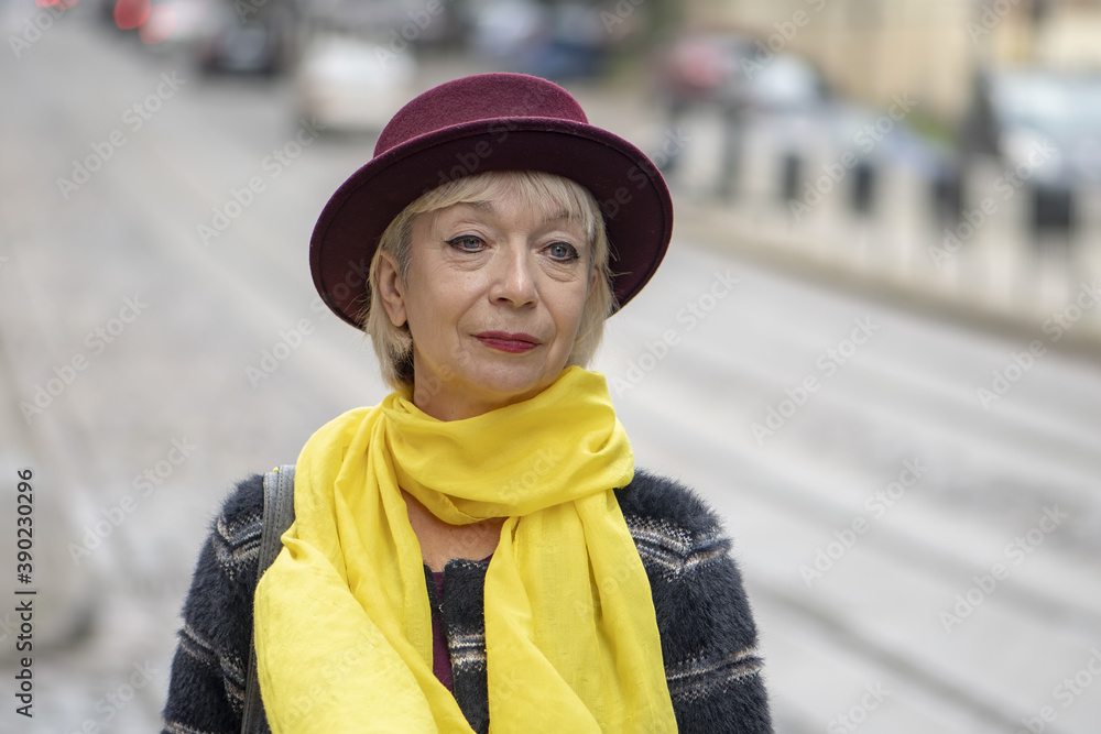 Street portrait of an elderly woman in a fashionable hat and yellow scarf on the background of a European urban landscape, medium plan, selective focus. Concept: excellent health in retirement,