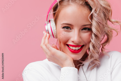 Close-up portrait on pink isolated background, girl with beautiful eyes and bright pink lips, listening to music through her headphones and sincerely smiling in front of lens