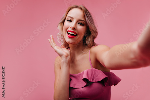 Pleased caucasian girl with blonde hair having fun in studio and taking picture of herself. Appealing white lady in dress making selfie