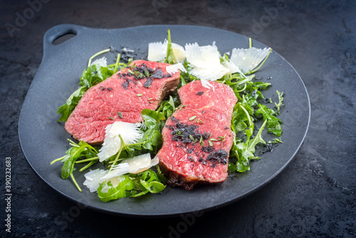 Modern style traditional fried New York strip steak with rucola and parmesan offered as close-up on a design plate